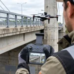 Osoba u rukavicama upravlja dronom koji lebdi pored mosta, uz prikaz mosta na ekranu upravljača